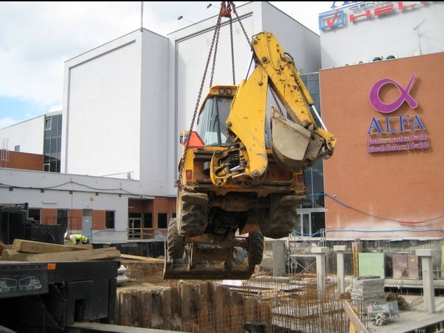 Żółta koparko-ładowarka podnoszona dźwigiem nad wykopem z widocznym zbrojeniem, na tle budynku Alfa z logo. Prace ziemne w przestrzeni miejskiej.