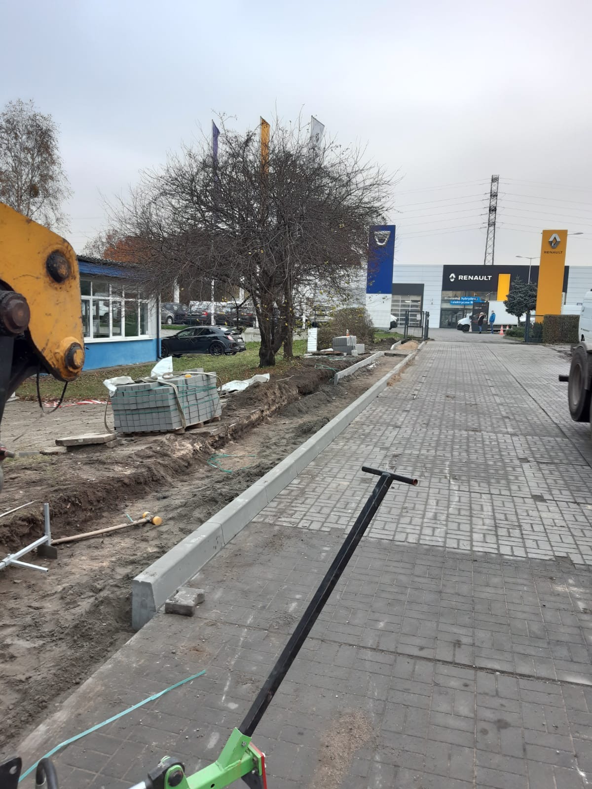 Układanie obrzeży chodnika z kostki brukowej przy salonie samochodowym. Widoczny fragment koparki i narzędzia. Obrzeża częściowo ułożone, kostka przygotowana.