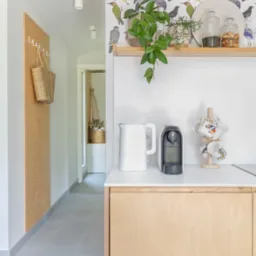 Wnętrze domu z elementami wykończenia z jasnej sklejki: szafka kuchenna, panel z wieszakami w przedpokoju, tapeta w ptaki, ekspres do kawy i czajnik na blacie.