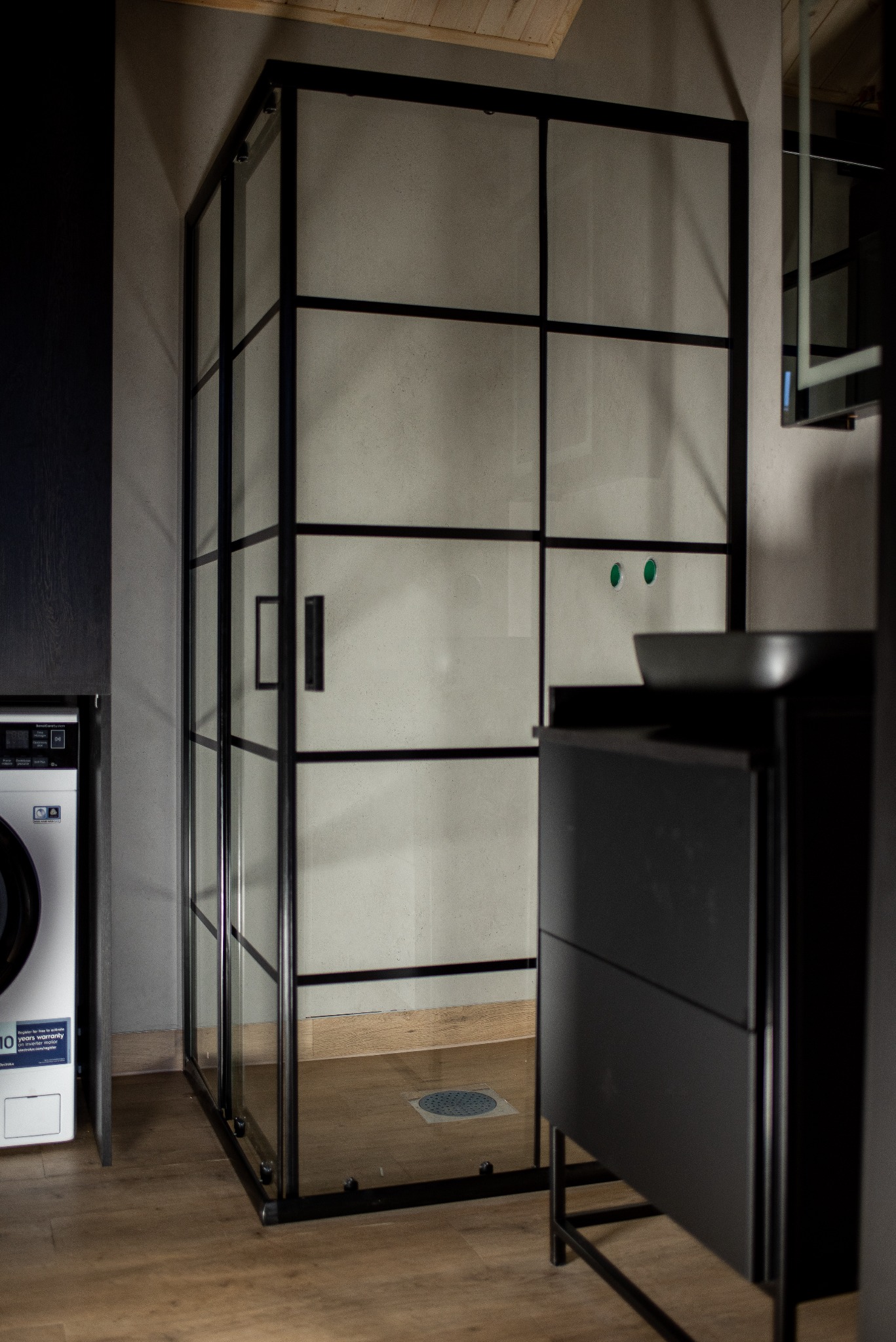 Nowoczesna kabina prysznicowa z czarnymi profilami i szklanymi ściankami, widoczna umywalka nablatowa i pralka w tle, całość w stonowanej kolorystyce.