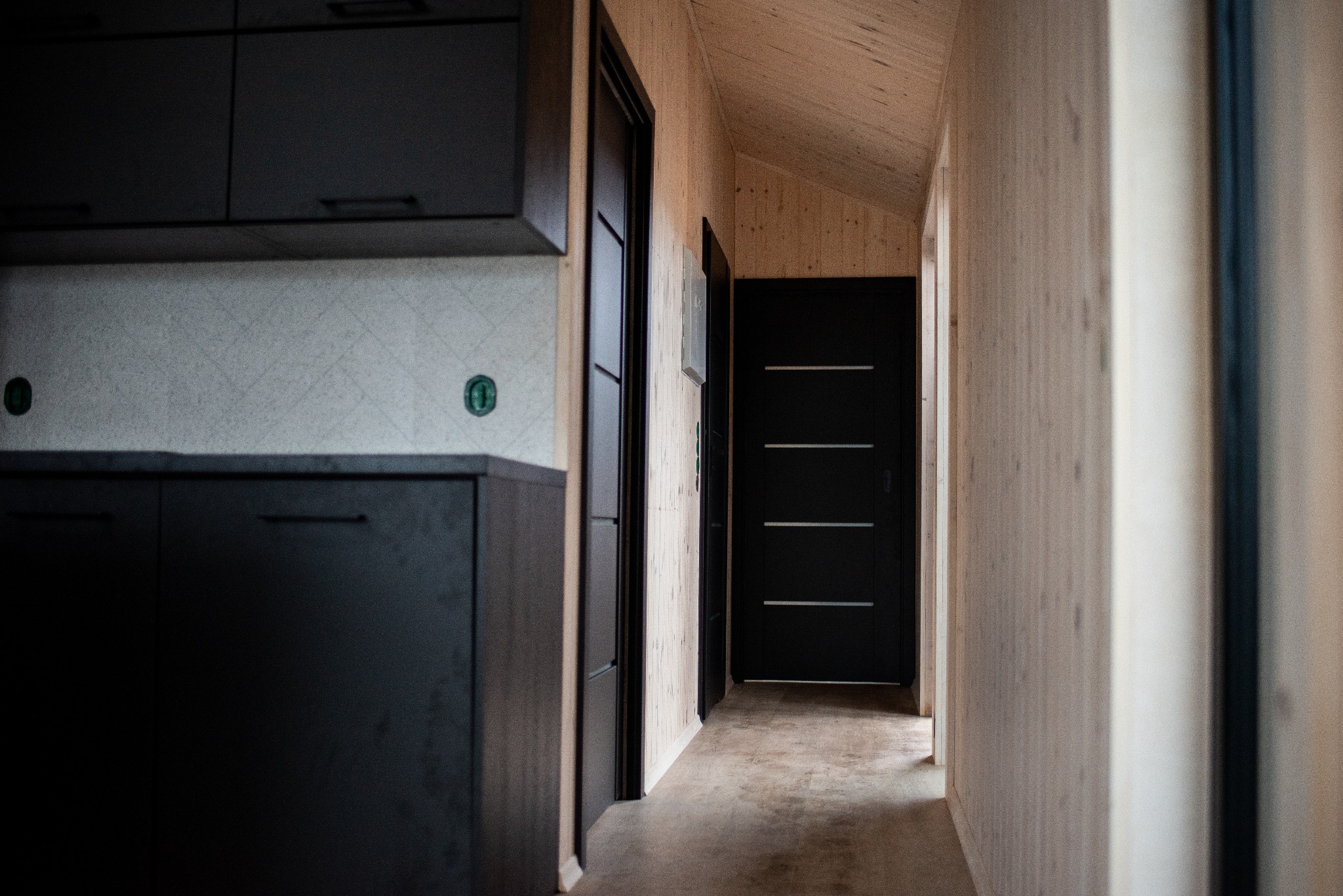 Wnętrze domu z drewnianymi ścianami i sufitem, czarne drzwi z poziomymi zdobieniami, fragment zabudowy kuchennej w ciemnym kolorze.