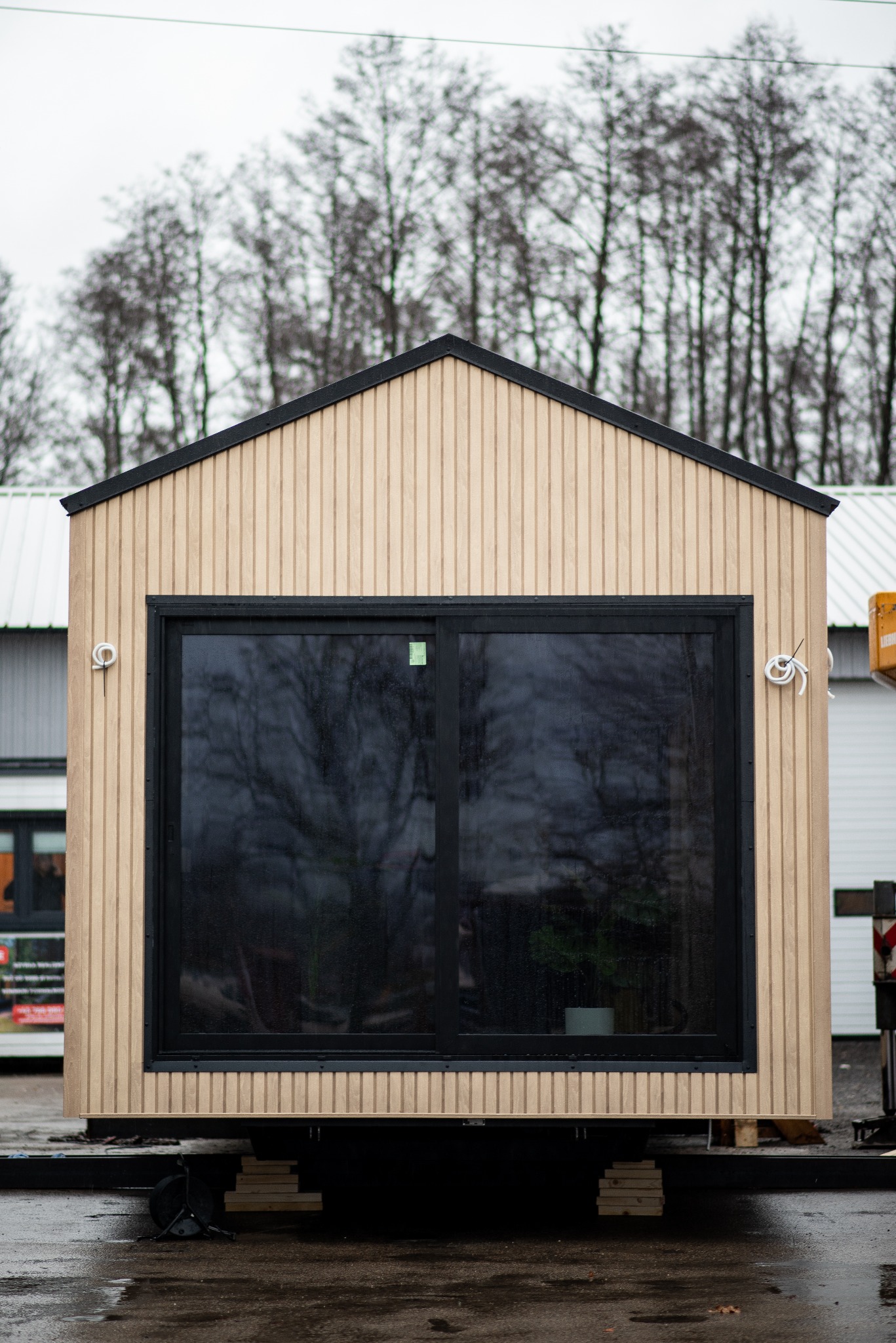 Mały, gotowy budynek z drewnianą elewacją, dużym panoramicznym oknem i czarnym obramowaniem dachu, umieszczony na platformie transportowej, na tle drzew bez liści.