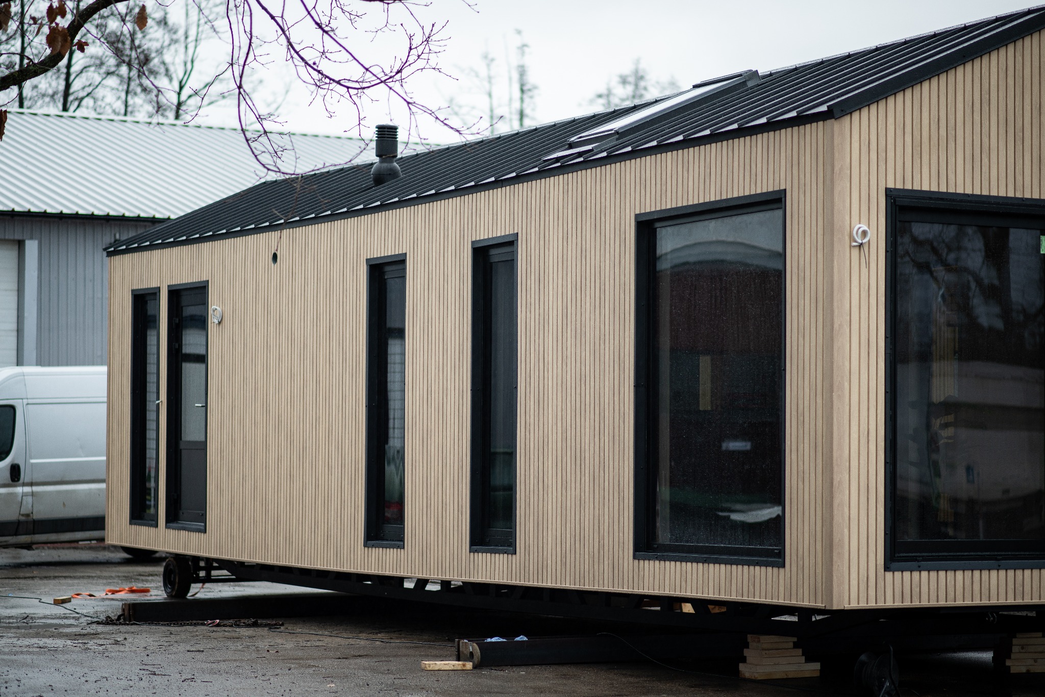 Przenośny dom z jasną, pionowo ułożoną okładziną drewnianą, czarną blachodachówką i czarnymi ramami okiennymi, ustawiony na metalowej konstrukcji z kołami, z widocznym kominem i fragmentem białego...