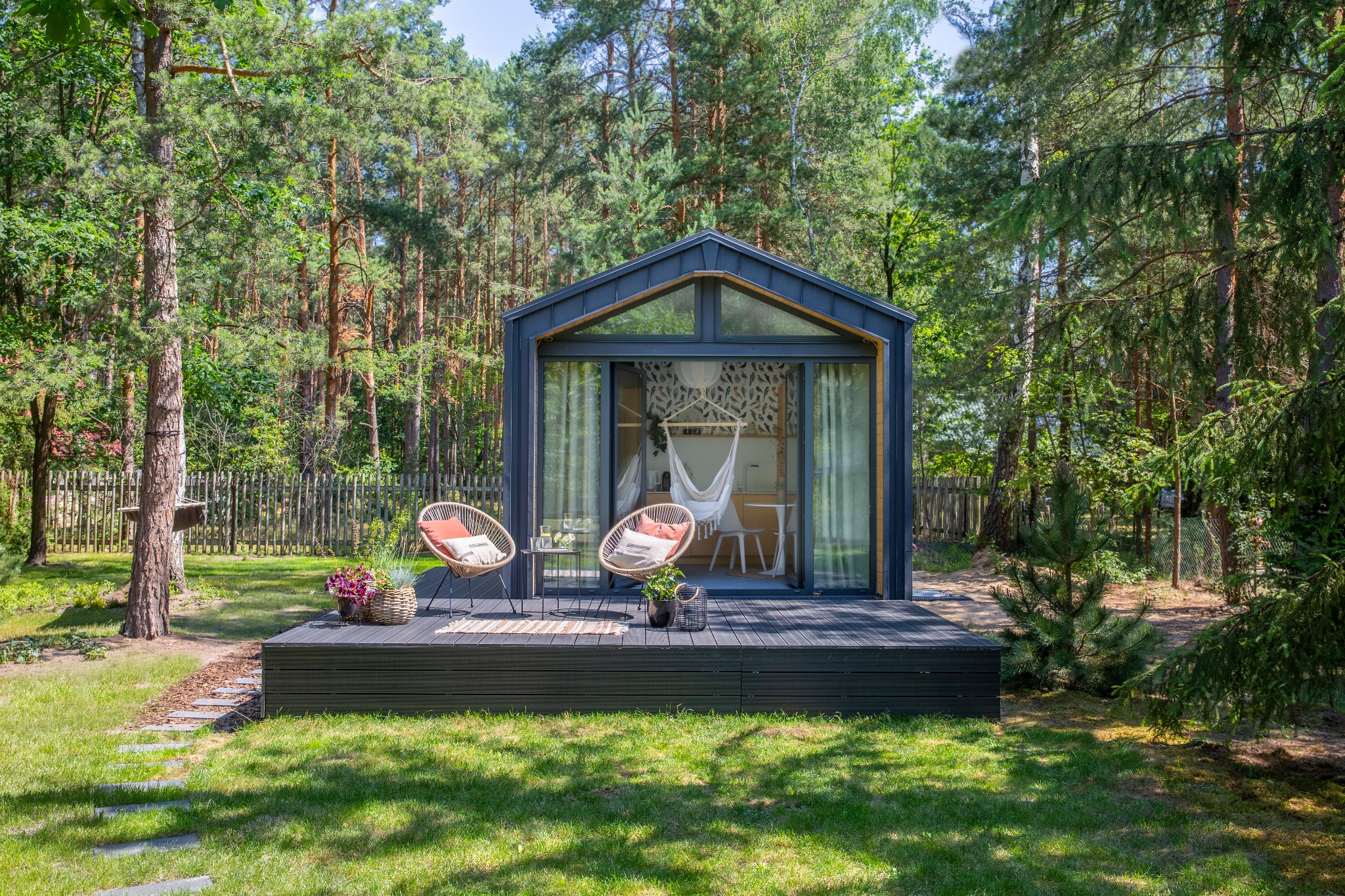 Nowoczesny, mały domek z dużymi przeszkleniami, tarasem z dwoma wiklinowymi fotelami i hamakiem wewnątrz, otoczony zielenią drzew.