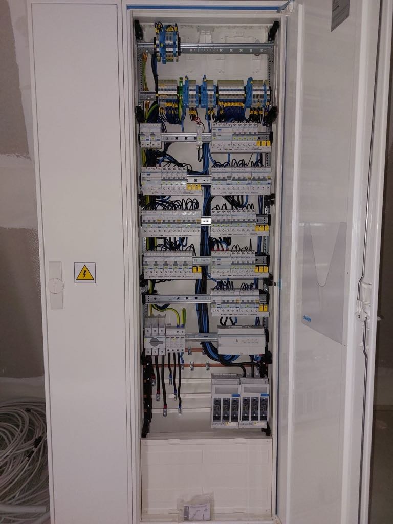 Otwarta szafa rozdzielcza z zamontowanymi bezpiecznikami, przewodami i elementami sterującymi, widoczny symbol ostrzegawczy na drzwiach szafy.