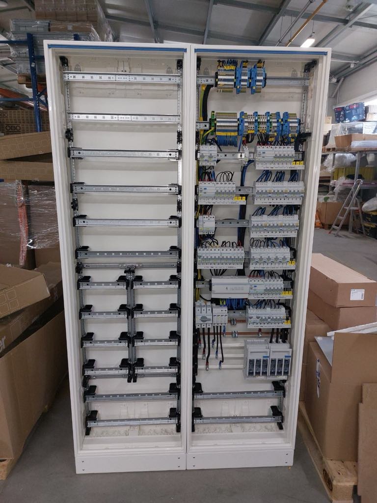 Rozdzielnica elektryczna w trakcie montażu, widoczne szyny DIN, przewody i zabezpieczenia, część szafy jeszcze pusta, otoczenie magazynowe.