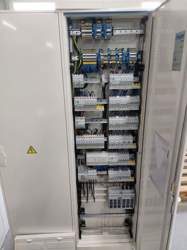 Otwarta szafa elektryczna z widocznymi bezpiecznikami, przewodami i oznaczeniami, żółty znak ostrzegawczy na drzwiach szafy.