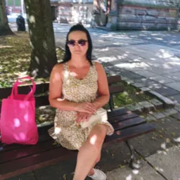 Kobieta w okularach przeciwsłonecznych siedzi na ławce w parku, obok różowa torba na zakupy, w tle budynek z graffiti.