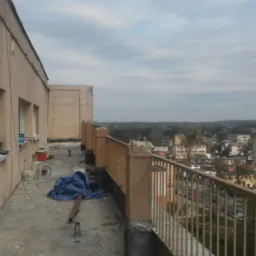 Elewacja budynku w trakcie remontu, widoczne ślady po tynkowaniu, farby, narzędzia i zabezpieczenia na balkonie z widokiem na miasto.