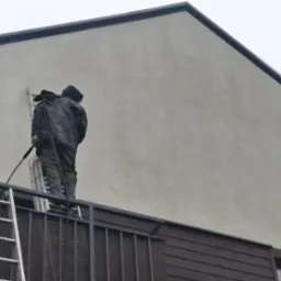 Pracownik w ubraniu roboczym myje elewację budynku z balkonu za pomocą myjki ciśnieniowej, stojąc na drabinie opierającej się o balustradę.