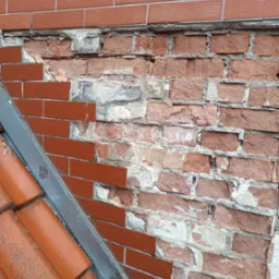 Fragment uszkodzonej, ceglanej elewacji z widocznymi ubytkami i fragmentem dachu pokrytego czerwoną dachówką, ukazujący potrzebę renowacji.