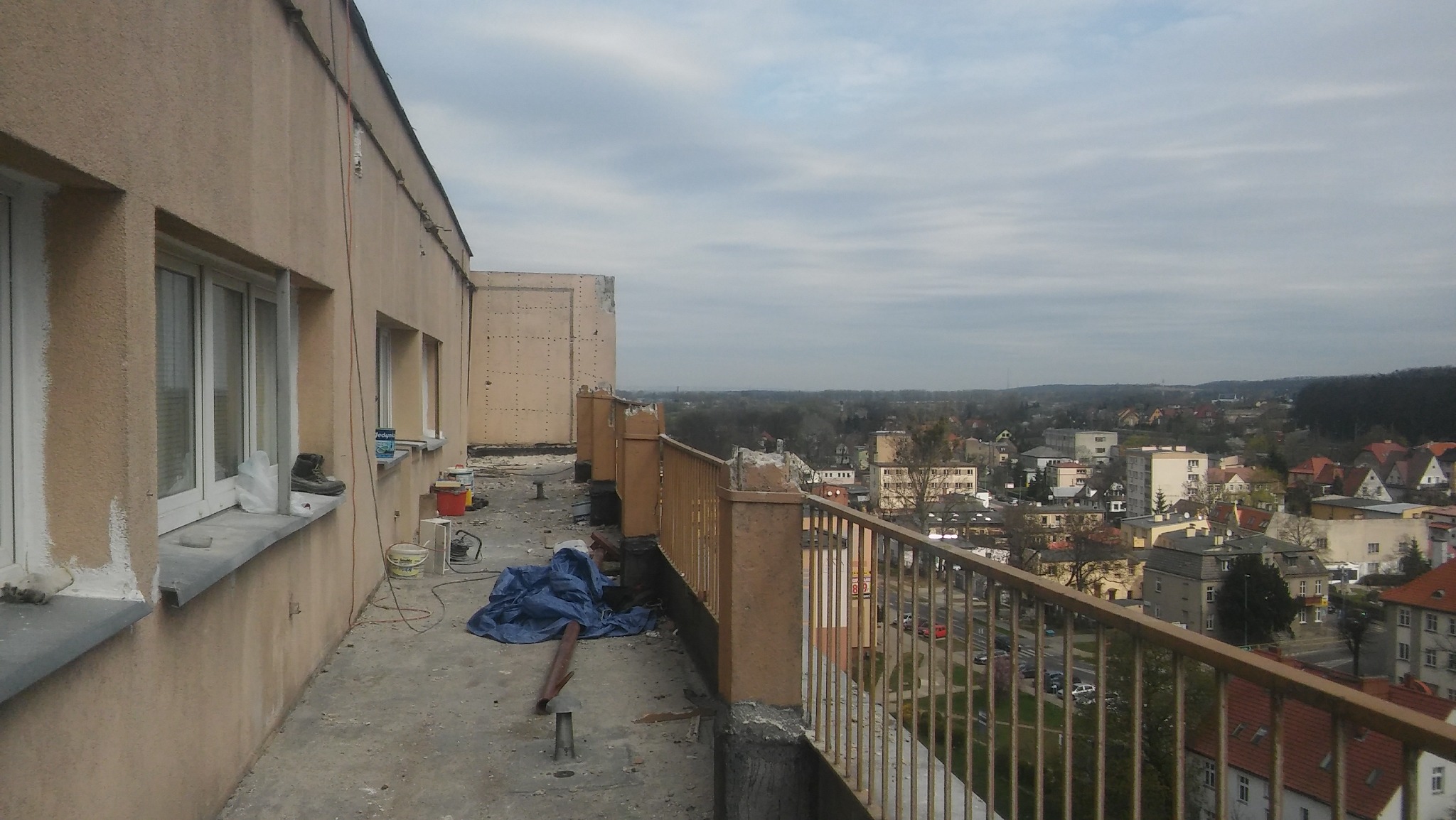 Elewacja budynku w trakcie remontu, widoczne ślady po tynkowaniu, farby, narzędzia i zabezpieczenia na balkonie z widokiem na miasto.