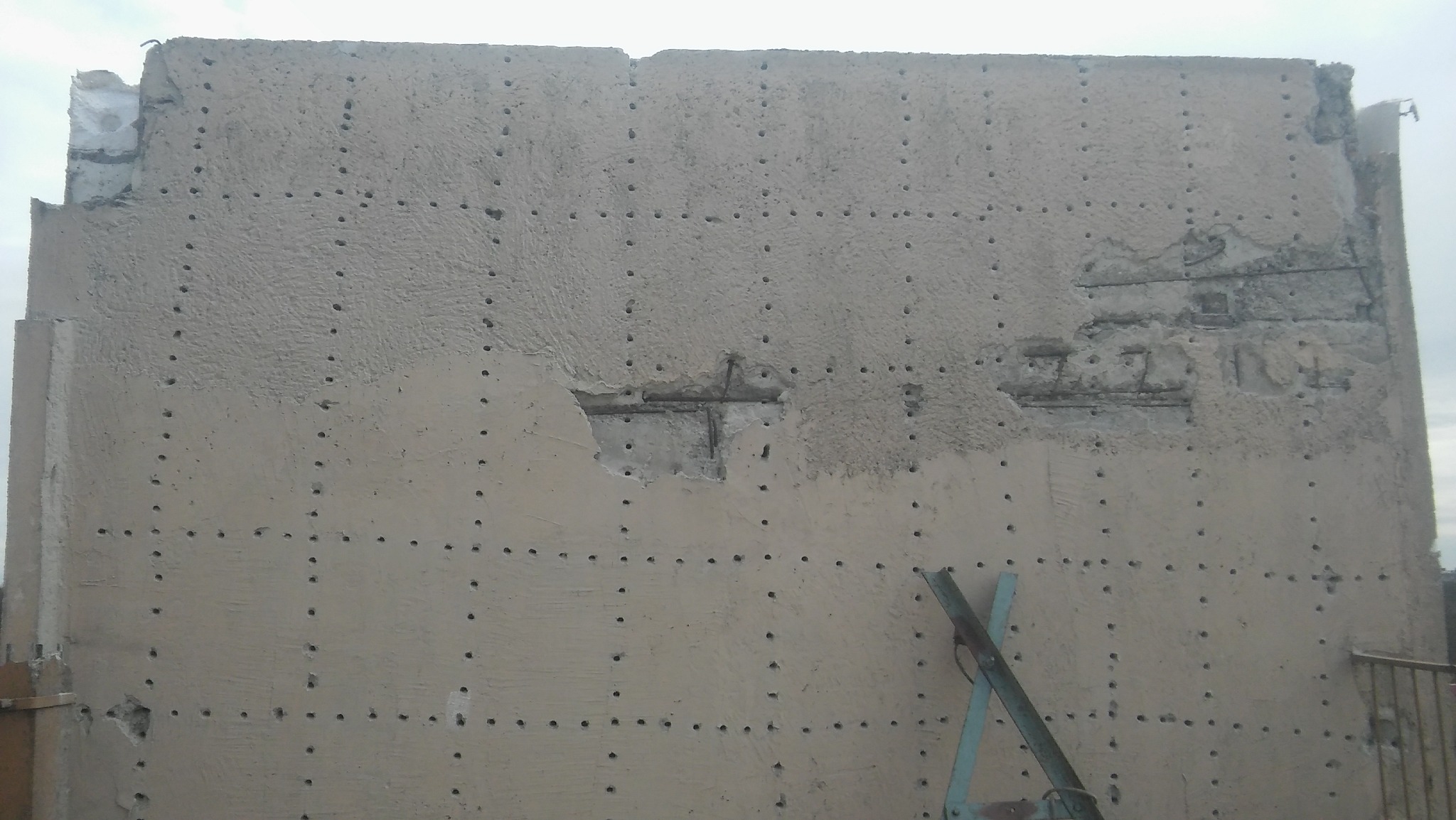 Uszkodzona elewacja budynku z widocznymi otworami i fragmentami odsłoniętej konstrukcji, częściowo naprawiona, z metalowym stojakiem opartym o ścianę.