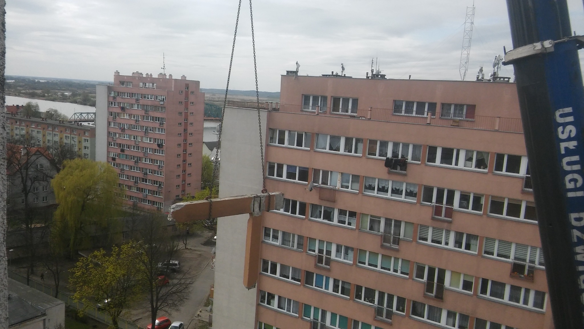 Fragment elewacji bloku mieszkalnego podnoszony przez dźwig na łańcuchach, widok z góry na budynki mieszkalne i drzewa w tle.