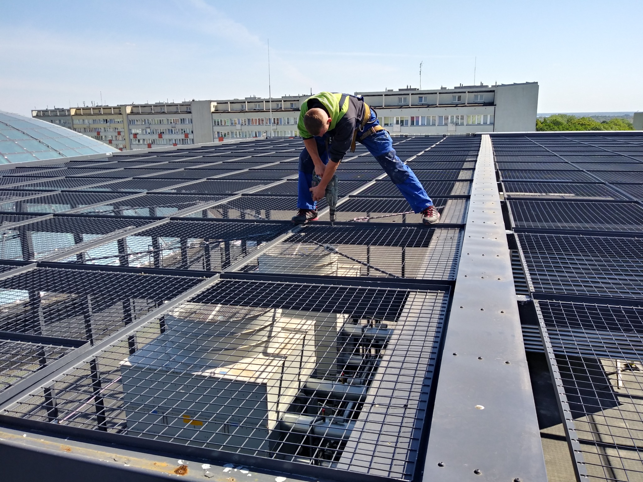 Pracownik w odblaskowej kamizelce wierci otwór w metalowej konstrukcji kratowej na dachu budynku z widokiem na odległe bloki mieszkalne.