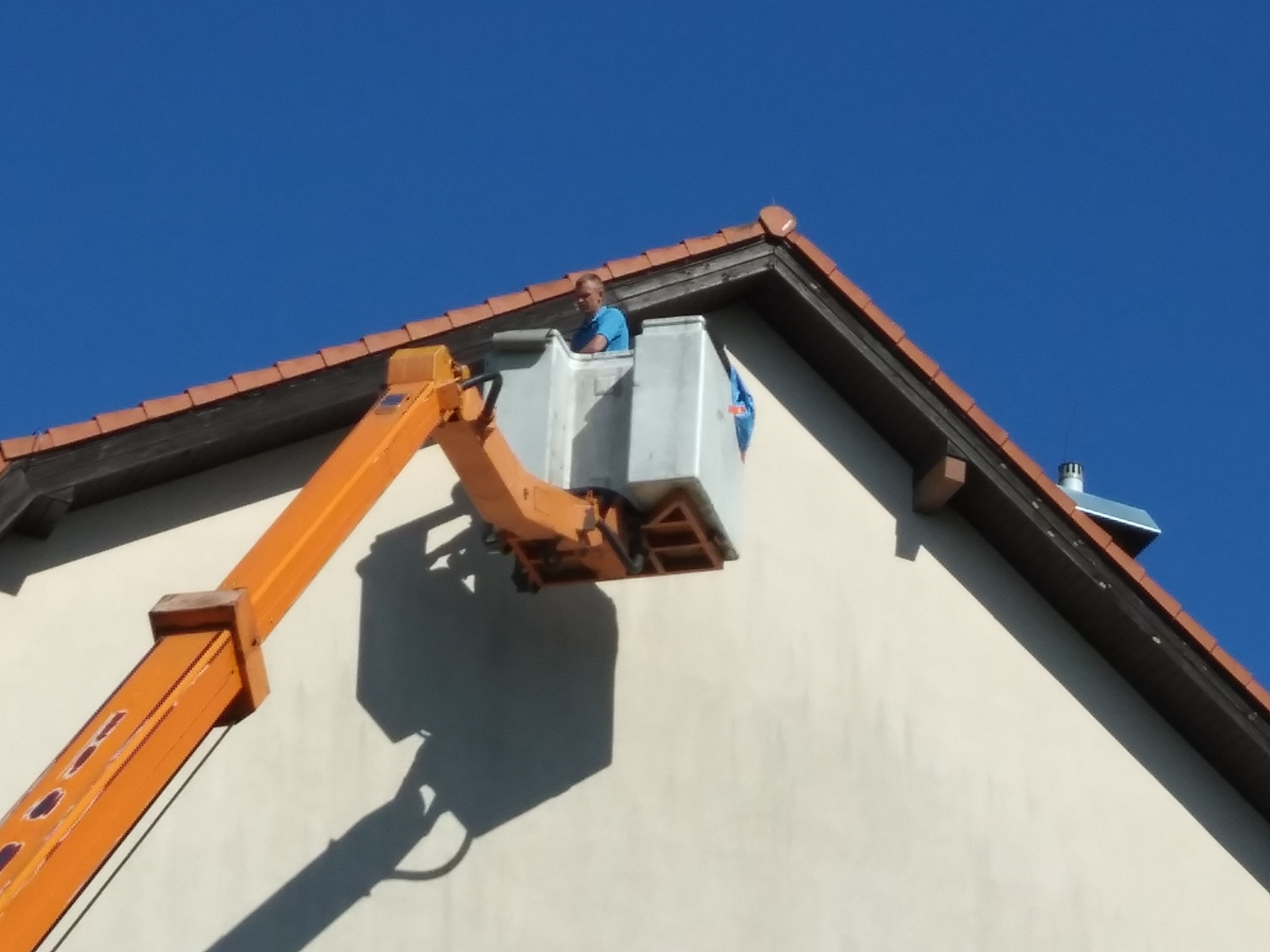 Pracownik w podnośniku koszowym na tle elewacji domu z brązową dachówką i błękitnego nieba.