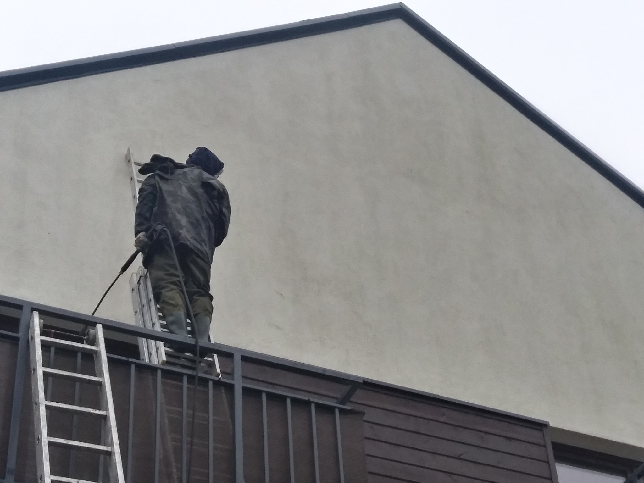 Pracownik w ubraniu roboczym myje elewację budynku z balkonu za pomocą myjki ciśnieniowej, stojąc na drabinie opierającej się o balustradę.
