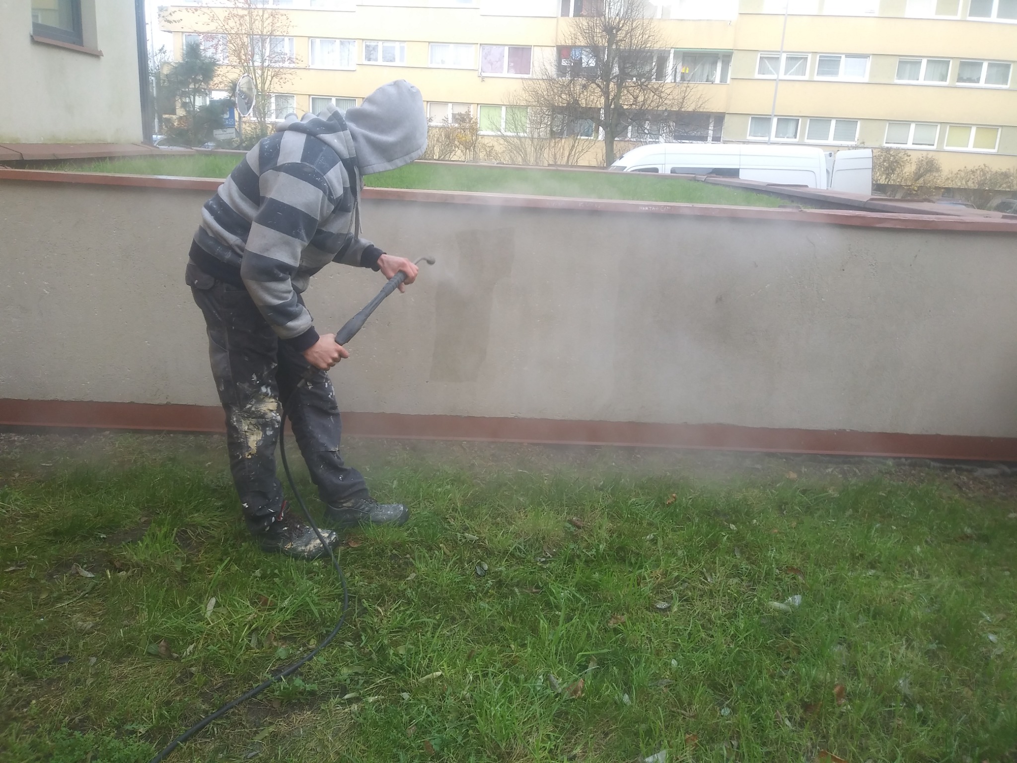 Pracownik w bluzie z kapturem czyści niską ścianę budynku myjką ciśnieniową, na trawniku widoczne ślady zabrudzeń.