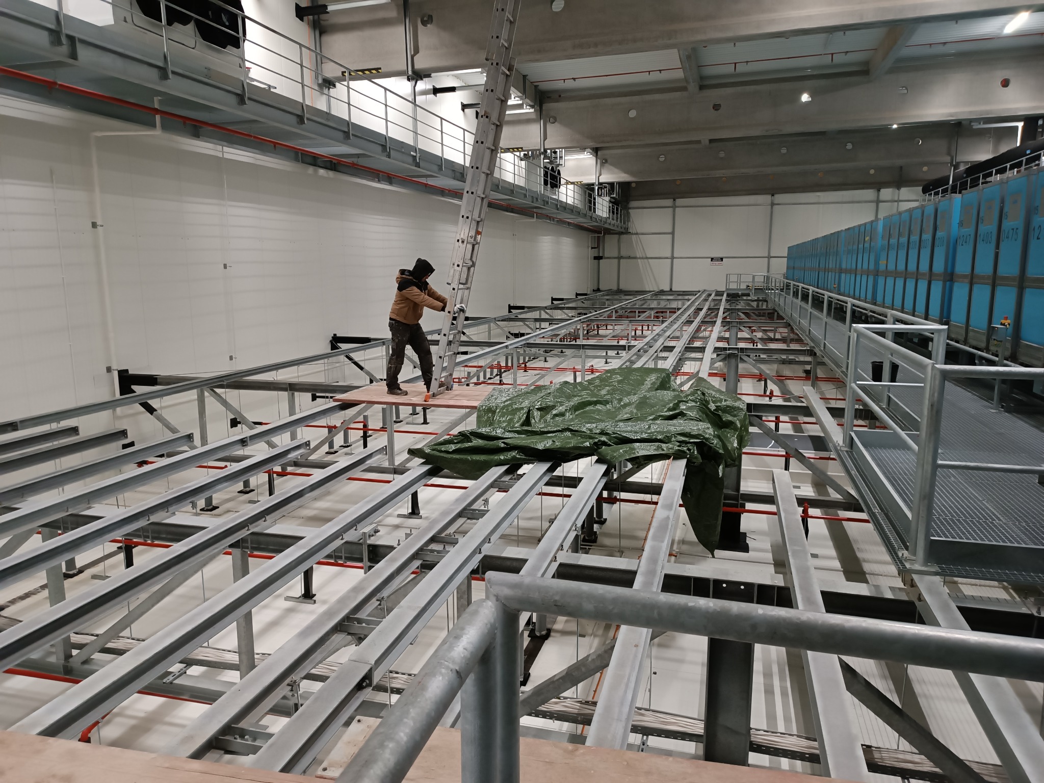 Pracownik w kombinezonie na platformie z metalowych belek w hali przemysłowej, w tle regały magazynowe i drabina oparta o konstrukcję.