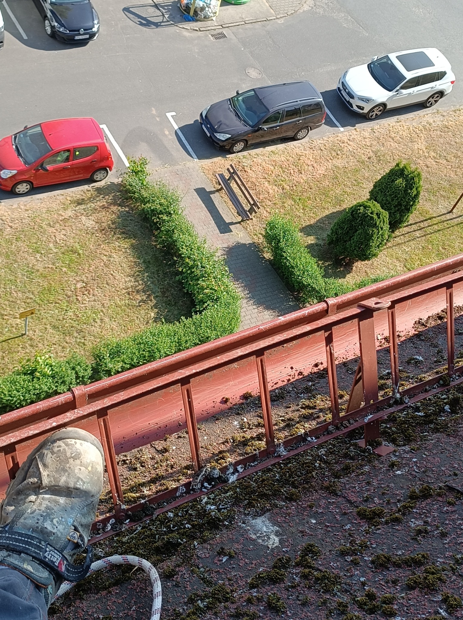 Stopa w zabrudzonym bucie roboczym na drabinie opierającej się o zarośnięty mchem dach, widok z góry na parking z samochodami i zielenią.