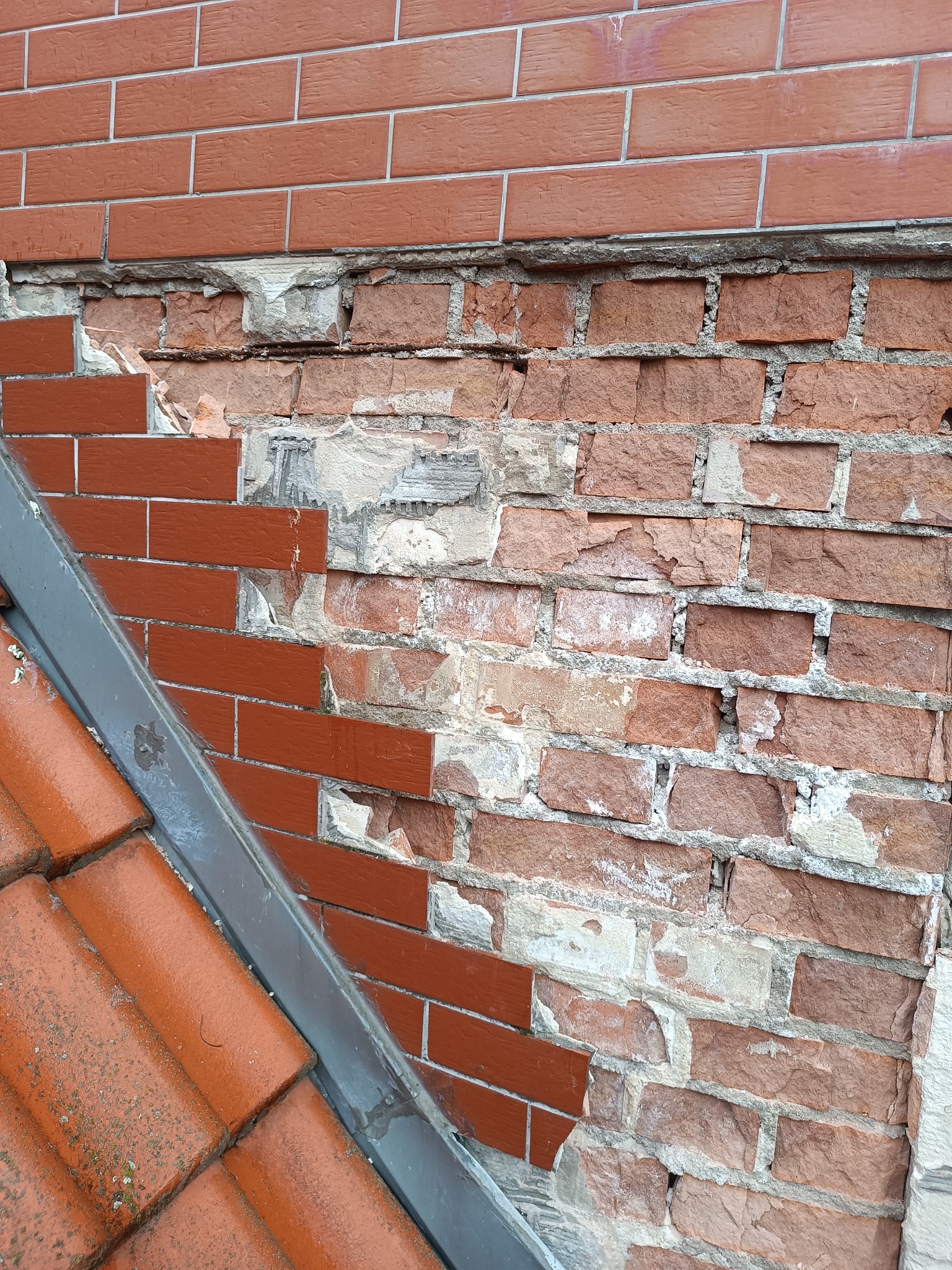 Fragment uszkodzonej, ceglanej elewacji z widocznymi ubytkami i fragmentem dachu pokrytego czerwoną dachówką, ukazujący potrzebę renowacji.