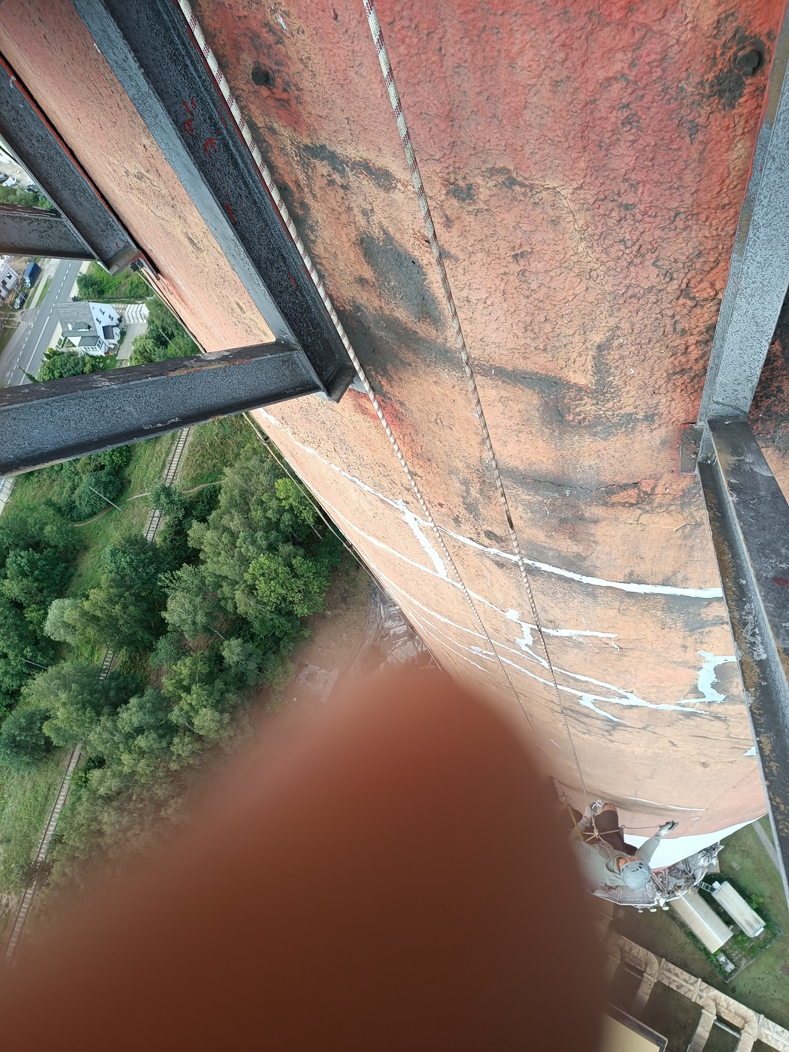 Pracownik na linie zabezpieczającej, wykonujący prace na wysokości na zewnątrz budynku, widoczne ślady wcześniejszych prac na elewacji i elementy konstrukcji stalowej.