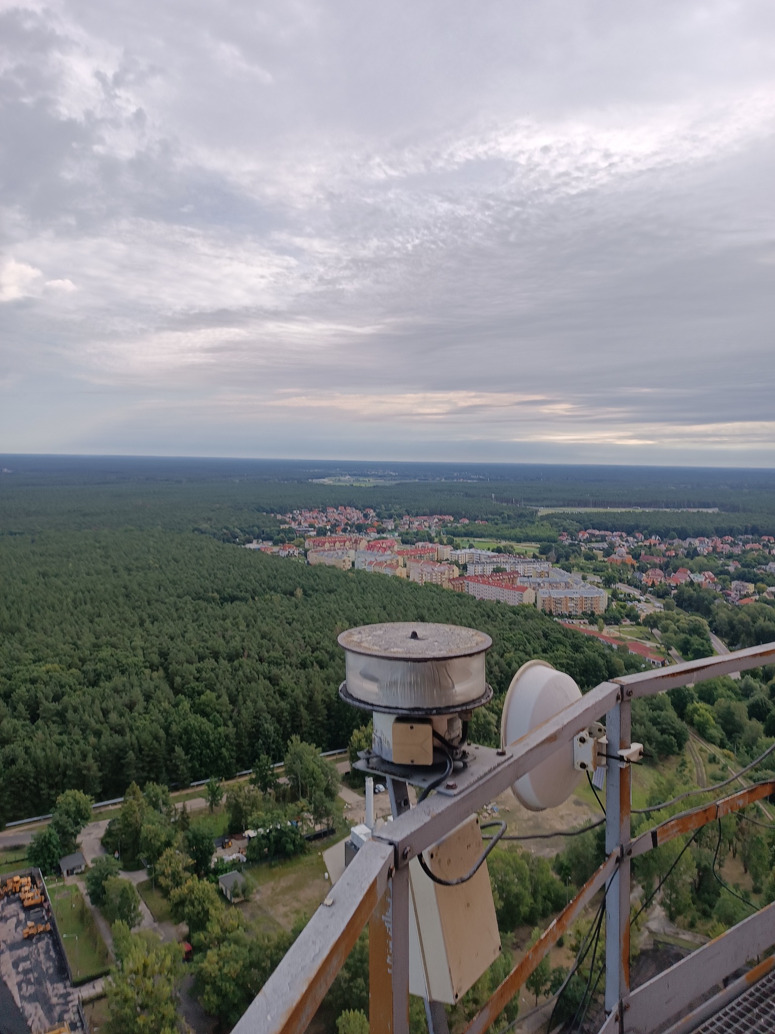 Widok z metalowej konstrukcji na panoramę miasta i lasu pod pochmurnym niebem; na pierwszym planie urządzenia telekomunikacyjne.