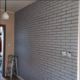 Ściana wewnętrzna pomalowana na szaro imitująca cegłę, z widocznymi gniazdkami elektrycznymi i drabiną w tle.