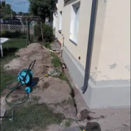 Ocieplenie fundamentów budynku w Choczewku, widoczny fragment elewacji z szarą, świeżo nałożoną warstwą izolacyjną, obok leży zwinięty wąż ogrodowy i narzędzia.