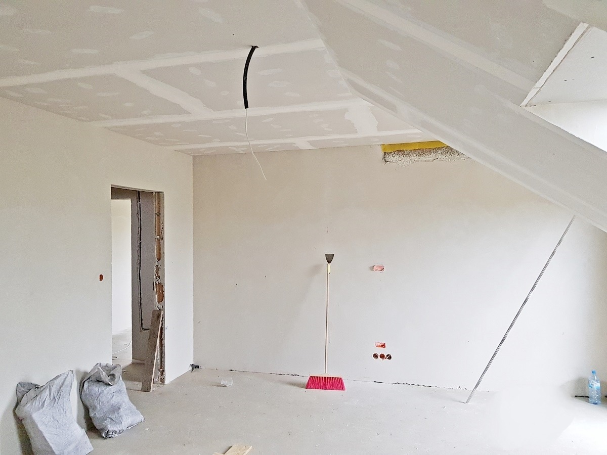 Poddasze w trakcie wykańczania: zamontowane płyty kartonowo-gipsowe na suficie, ściany przygotowane do dalszych prac, widoczne worki z materiałami, narzędzia (szpachelka, miotła) oraz wystające...