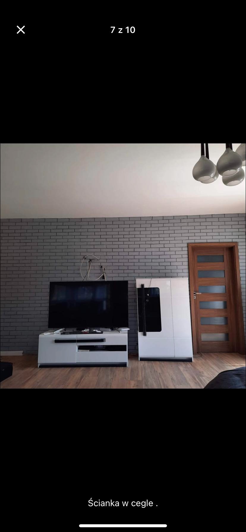 Pokój z szarą ścianą wyłożoną cegłą, białe meble pod telewizorem i drewniane drzwi z przeszkleniami.