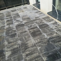 Dawid Barczyk - Szary taras wyłożony kwadratową kostką o nieregularnym wzorze, przylegający do białej ściany z czarnymi ramami okiennymi i balustradą.