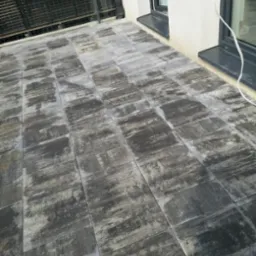 Szary taras wyłożony kwadratową kostką o nieregularnym wzorze, przylegający do białej ściany z czarnymi ramami okiennymi i balustradą.