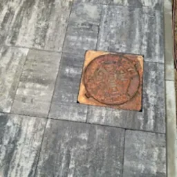 Szare, prostokątne płyty chodnikowe ułożone wokół okrągłej, żeliwnej studzienki kanalizacyjnej z widocznym wzorem i napisem.