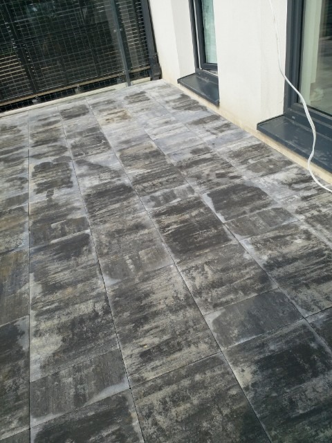 Szary taras wyłożony kwadratową kostką o nieregularnym wzorze, przylegający do białej ściany z czarnymi ramami okiennymi i balustradą.