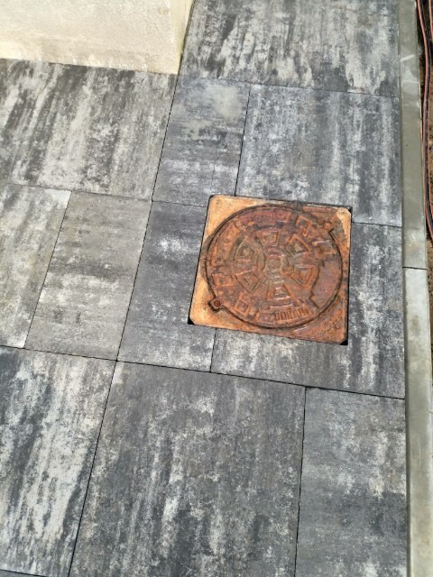 Szare, prostokątne płyty chodnikowe ułożone wokół okrągłej, żeliwnej studzienki kanalizacyjnej z widocznym wzorem i napisem.