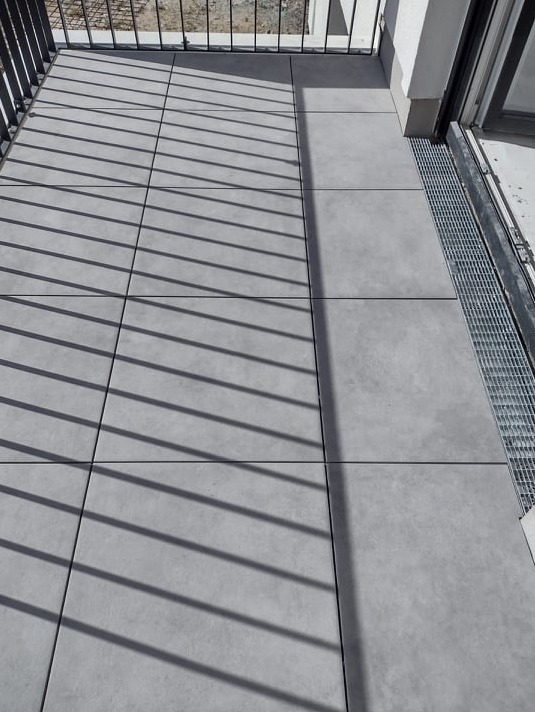 Szare płytki tarasowe ułożone na balkonie, cień barierki tworzy geometryczny wzór na powierzchni.
