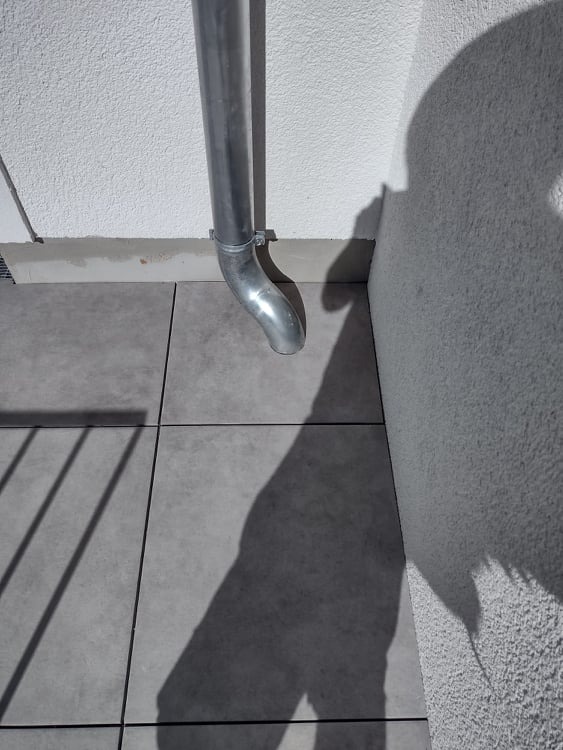 Szara płytka tarasowa z widoczną rynną spustową i cieniem barierki balkonowej na powierzchni.