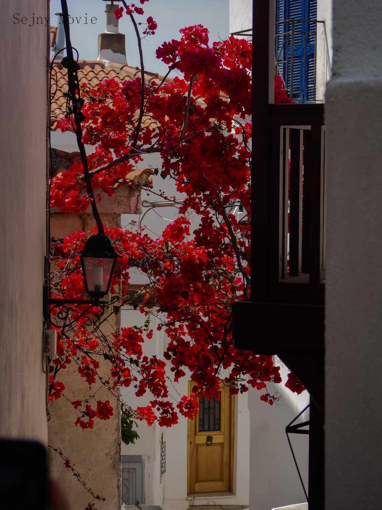 Uliczka w śródziemnomorskim miasteczku z kwitnącą bugenwillą, widok zza rogu budynku na białe ściany, drewniane drzwi, balkon z niebieskimi okiennicami i ozdobną latarnię.
