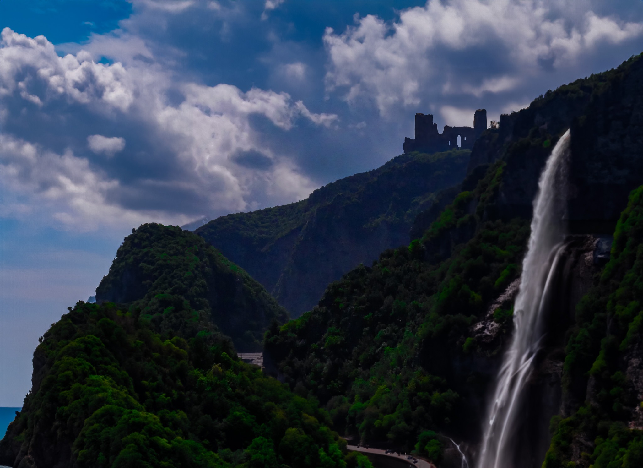 Malowniczy krajobraz z wodospadem spływającym po zboczu góry porośniętej bujną zielenią, na szczycie której widać ruiny zamku na tle pochmurnego nieba.
