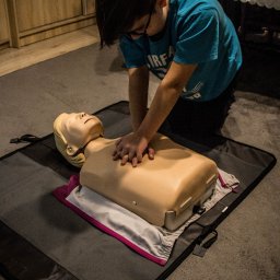 BIŁAS-MED Szkolenia z pierwszej pomocy - Młody mężczyzna w okularach klęczy na szarej macie i wykonuje resuscytację krążeniowo-oddechową na manekinie z blond włosami, umieszczonym na złożonych ubraniach, w pomieszczeniu z drewnianą szafką...