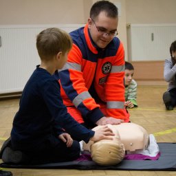 BIŁAS-MED Szkolenia z pierwszej pomocy - Instruktor w pomarańczowej kurtce z logiem ratownictwa medycznego uczy chłopca wykonywania resuscytacji na manekinie podczas kursu pierwszej pomocy, obserwują inni uczestnicy.