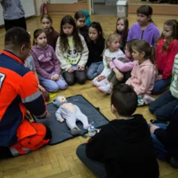 Grupa dzieci siedzi na podłodze w sali gimnastycznej, przodem do ratownika medycznego w pomarańczowym uniformie, który demonstruje resuscytację na manekinie niemowlęcia leżącym na szarej macie.