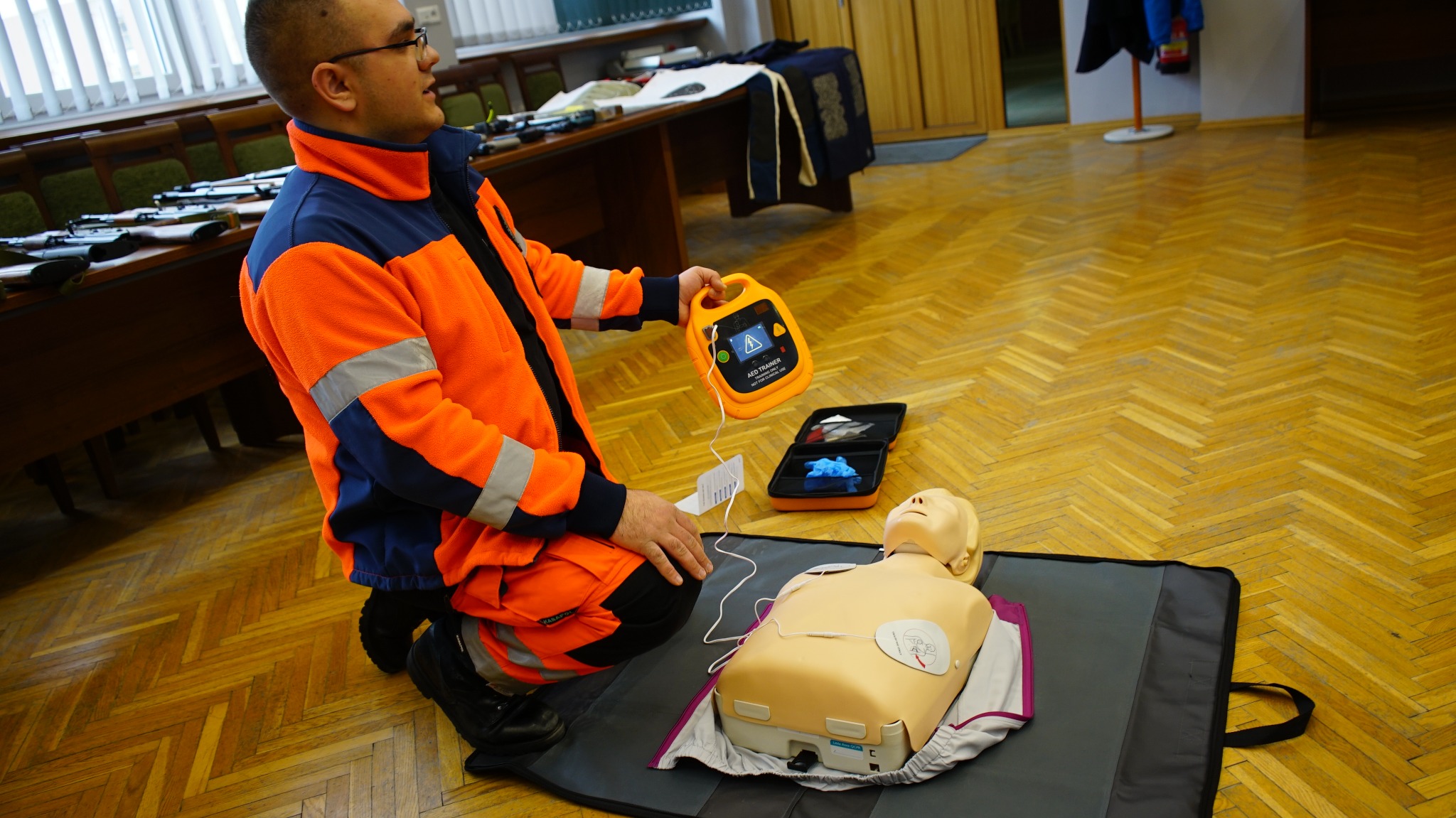 Instruktor w odzieży roboczej demonstruje użycie defibrylatora AED na manekinie podczas zajęć praktycznych, na tle parkietu i stołu z atrapami broni.