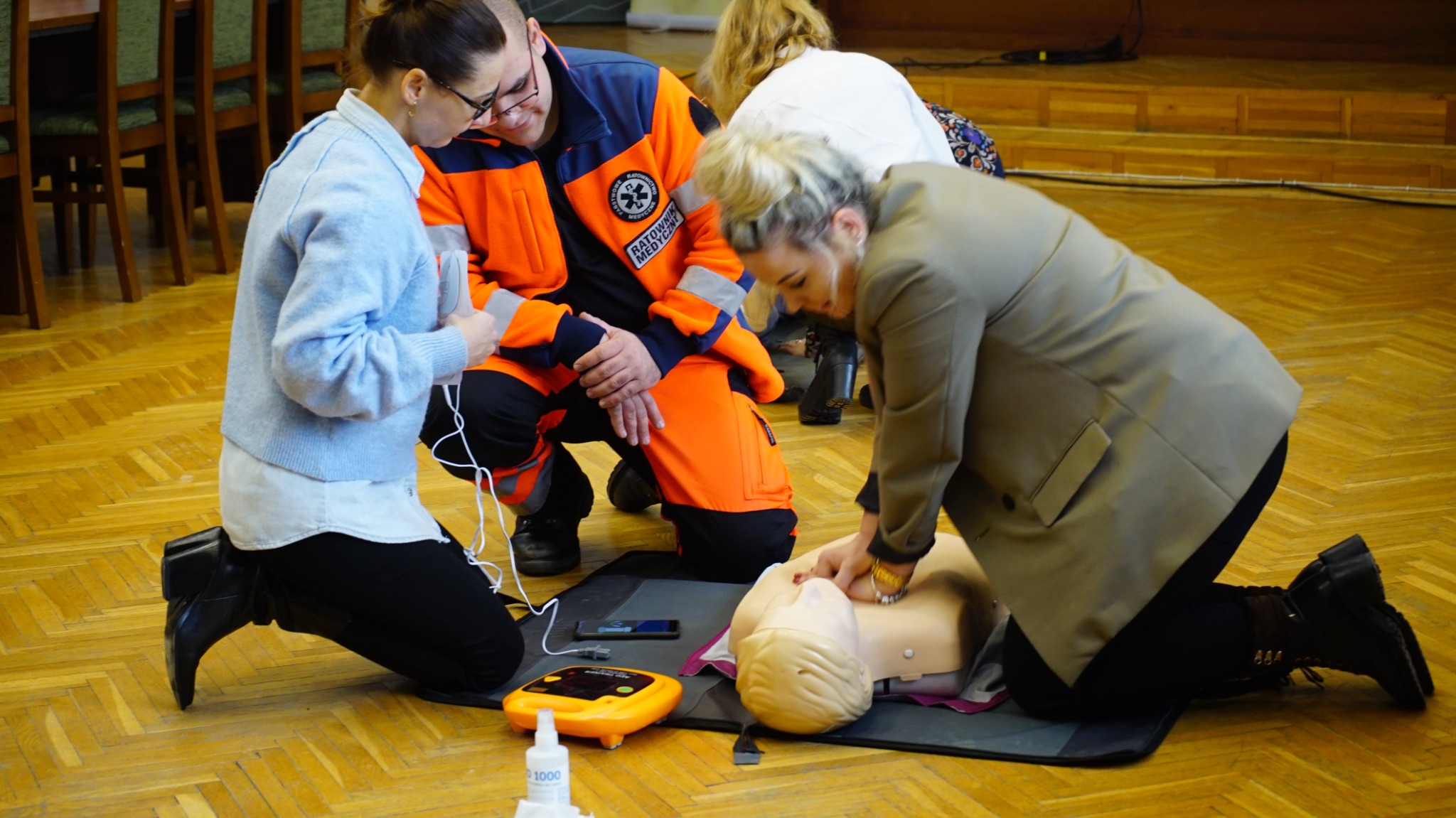 Ćwiczenia resuscytacji krążeniowo-oddechowej na fantomie, instruktor w pomarańczowym uniformie ratownika medycznego nadzoruje kursantkę w beżowym żakiecie, obok defibrylator i preparat...