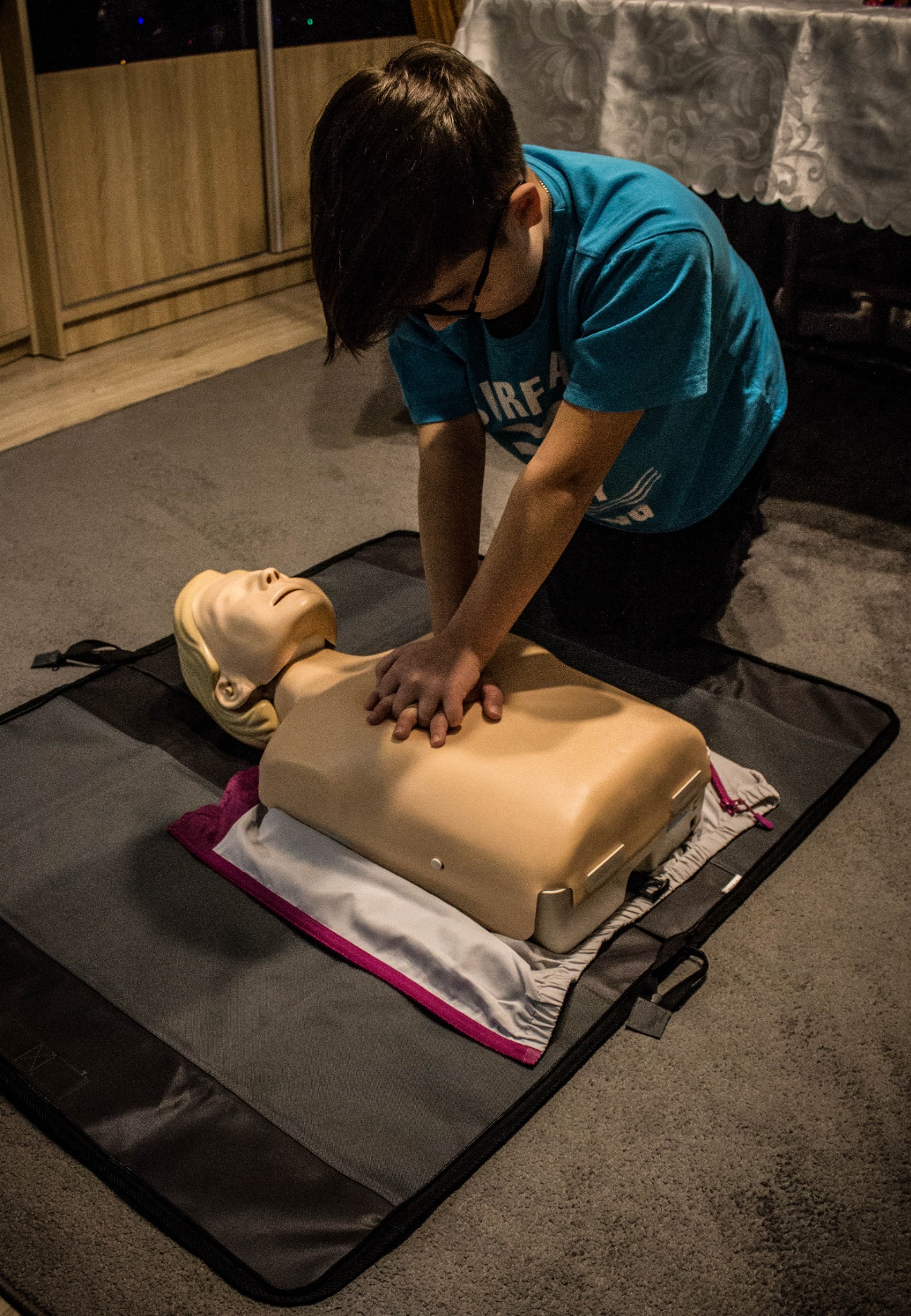 Młody mężczyzna w okularach klęczy na szarej macie i wykonuje resuscytację krążeniowo-oddechową na manekinie z blond włosami, umieszczonym na złożonych ubraniach, w pomieszczeniu z drewnianą szafką...