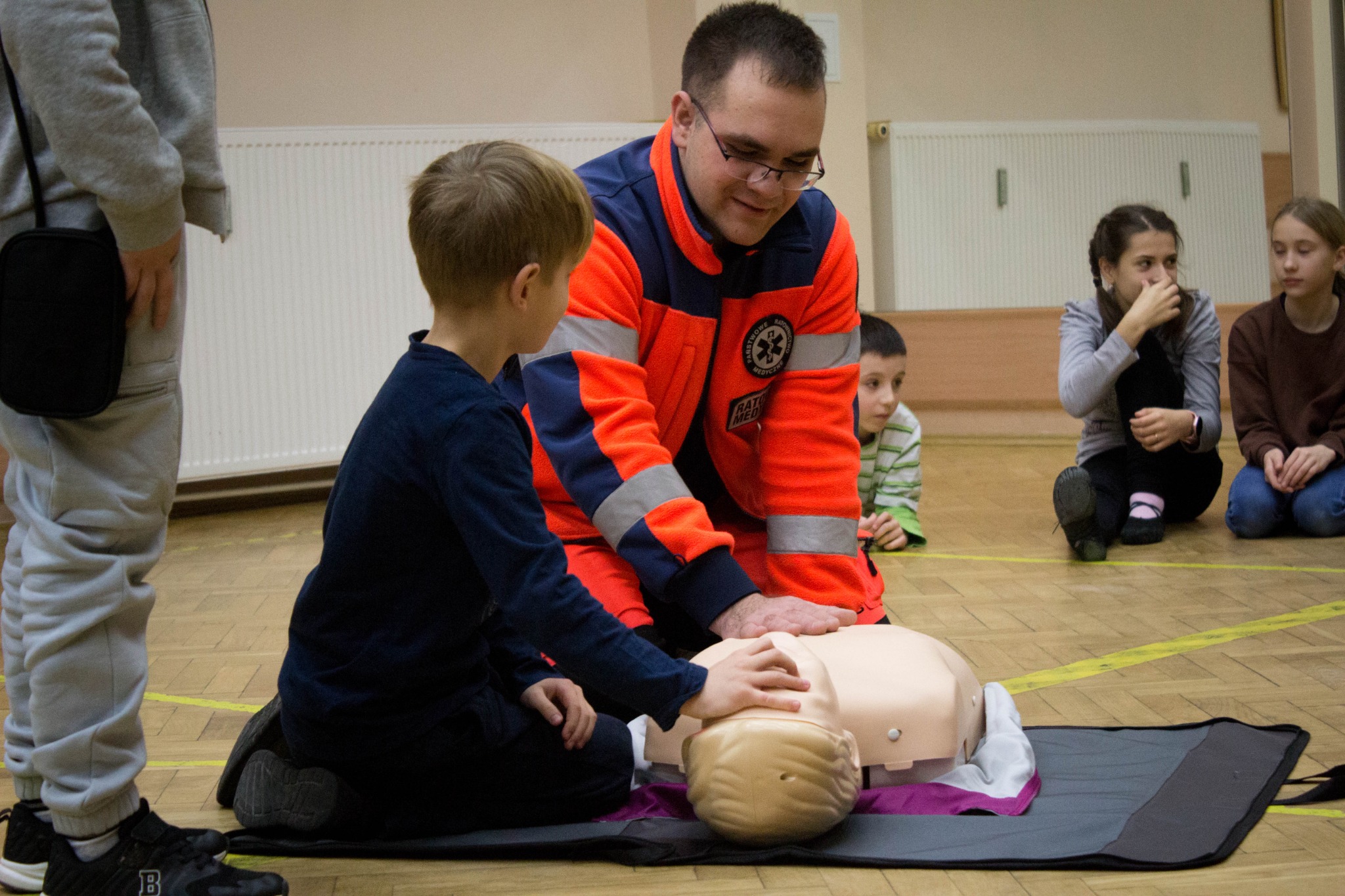 Instruktor w pomarańczowej kurtce z logiem ratownictwa medycznego uczy chłopca wykonywania resuscytacji na manekinie podczas kursu pierwszej pomocy, obserwują inni uczestnicy.