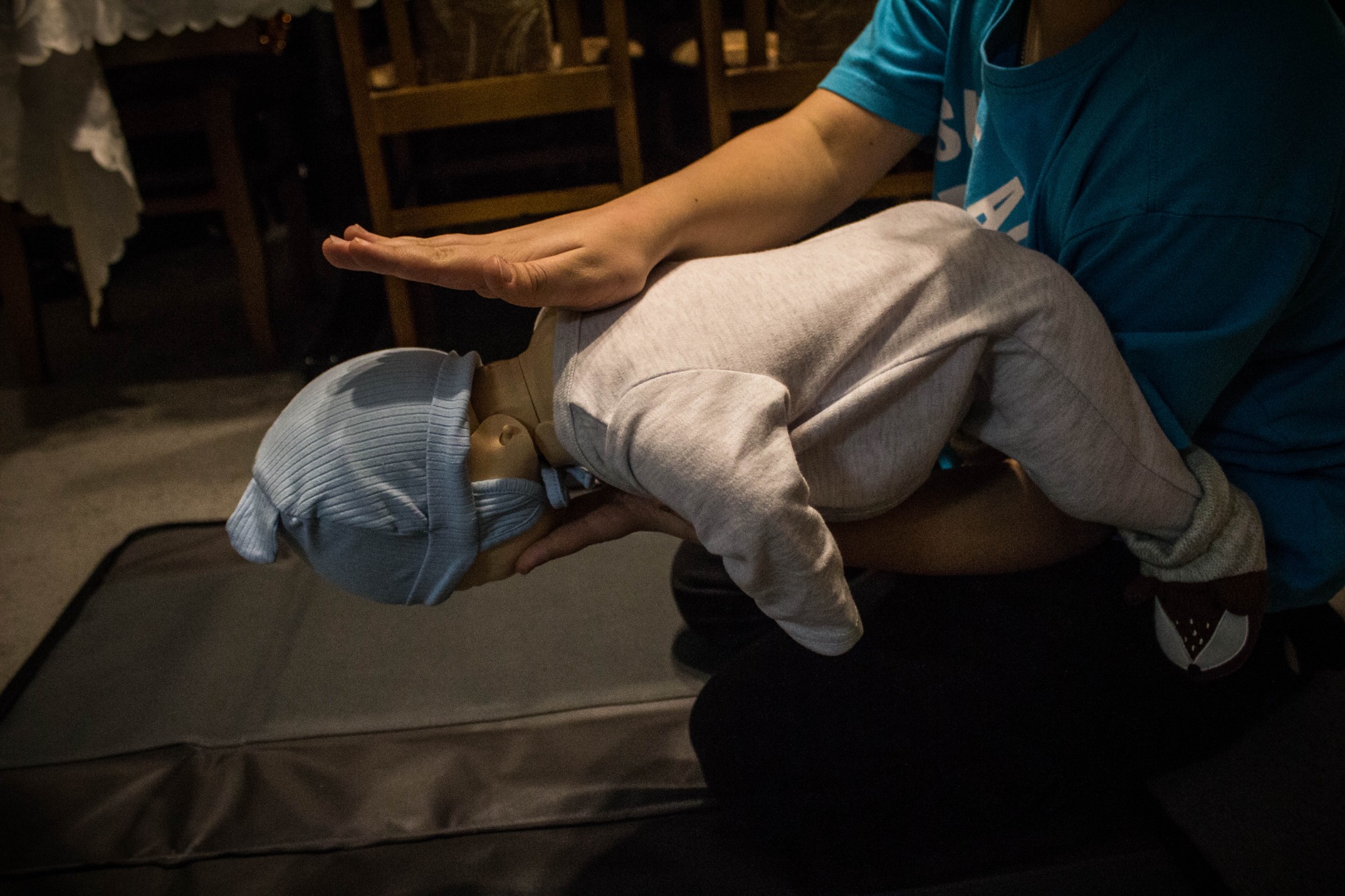 Instruktor demonstruje technikę resuscytacji niemowlęcia na manekinie, dłoń uniesiona nad klatką piersiową, manekin ubrany w jasny kombinezon i niebieską czapkę, w tle ciemne krzesła.