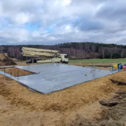 Wylany fundament pod budowę domu jednorodzinnego, widoczna pompa do betonu, zbrojenie i cegły na obrzeżach, otoczenie to teren budowy z piaskiem i lasem w tle, pochmurne niebo.