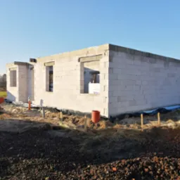 Wznoszone ściany parterowego domu z betonu komórkowego, widoczne otwory okienne, izolacja fundamentów i przygotowany teren budowy z paletami w tle.